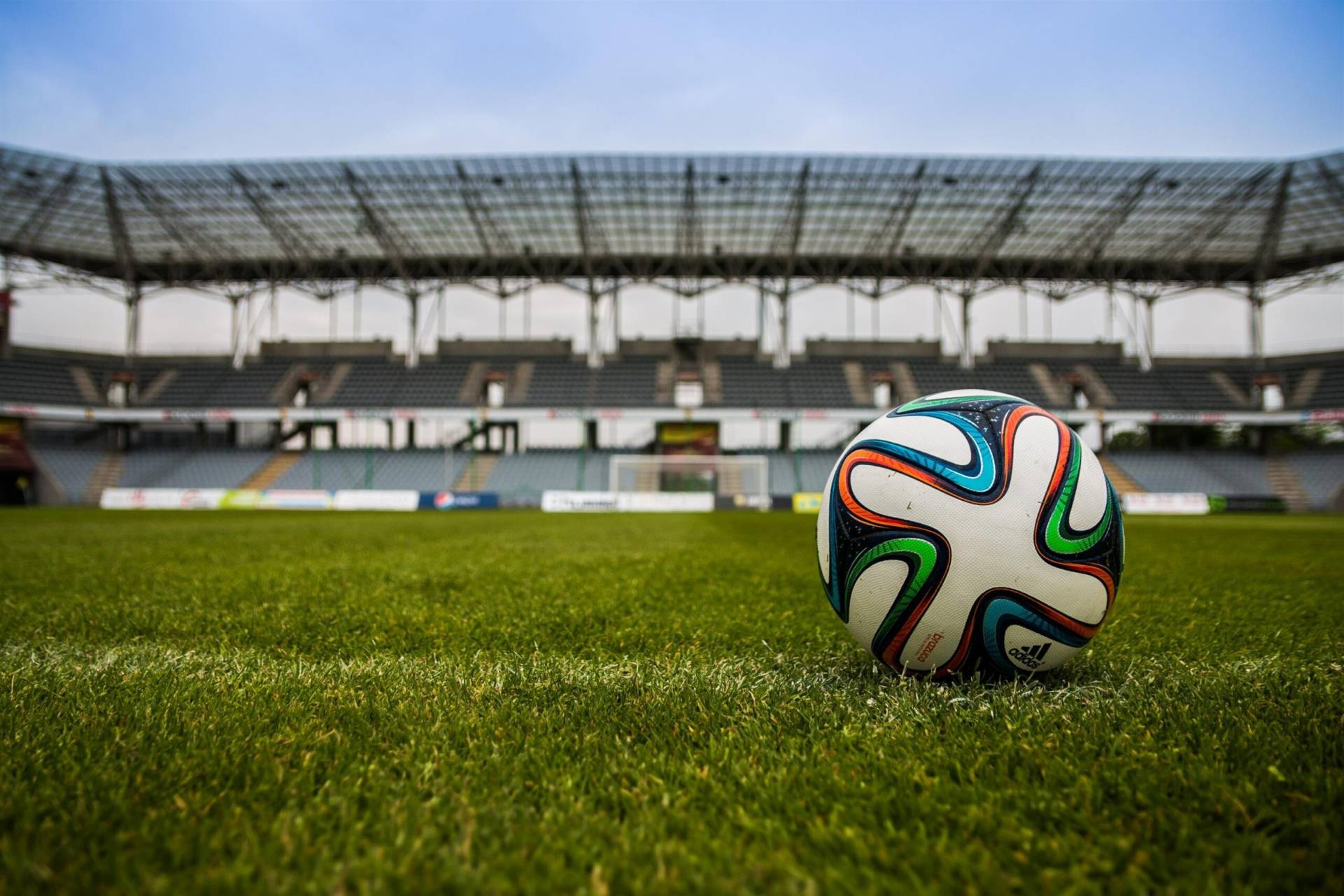 The photo is taken from the level of the ground. Photo of a football field, with green turf in the foreground along with a colourful football, and stadium and blue skies in the background.